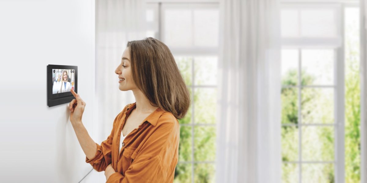 Young woman controlling home with a digital touch screen panel installed on the wall in the living room. Concept of a smart home and mobile application for managing smart devices at home
