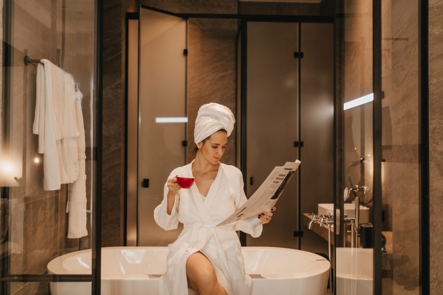 Woman in towel on her head and bathrobe reads latest news. Lady with cup of tea posing in bathroom